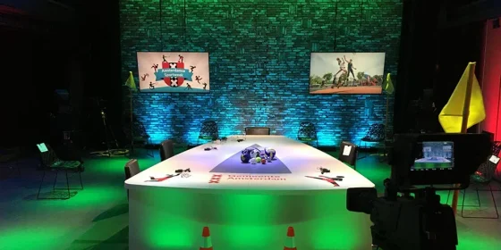 Studiodecor met groene verlichting, een tafel en schermen waarop de sprekers in beeld zijn.